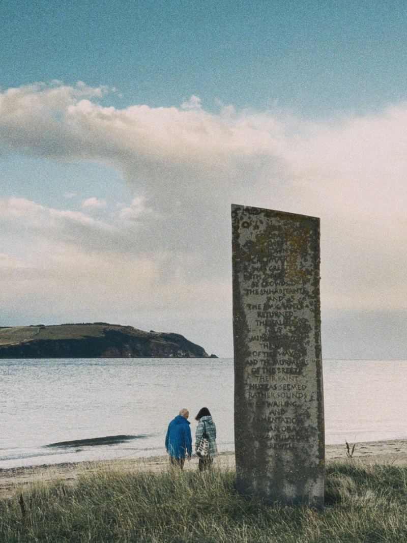 The Emigration Stone, a tall inscribed slab on the shore at Cromarty, with two people standing beside it